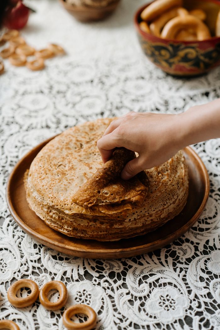 A hand picks a blin from a stack on a lace tablecloth, symbolizing Russian culinary tradition.